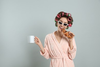 Beautiful young woman in bathrobe with hair curlers holding croissant and cup of drink on light grey background