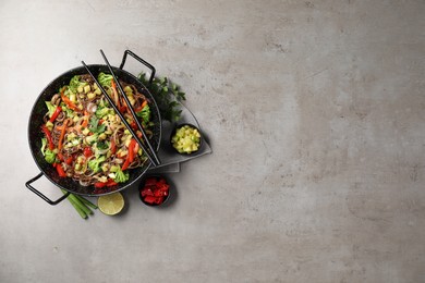 Photo of Stir-fry. Tasty noodles with meat in wok, chopsticks and ingredients on grey textured table, flat lay. Space for text