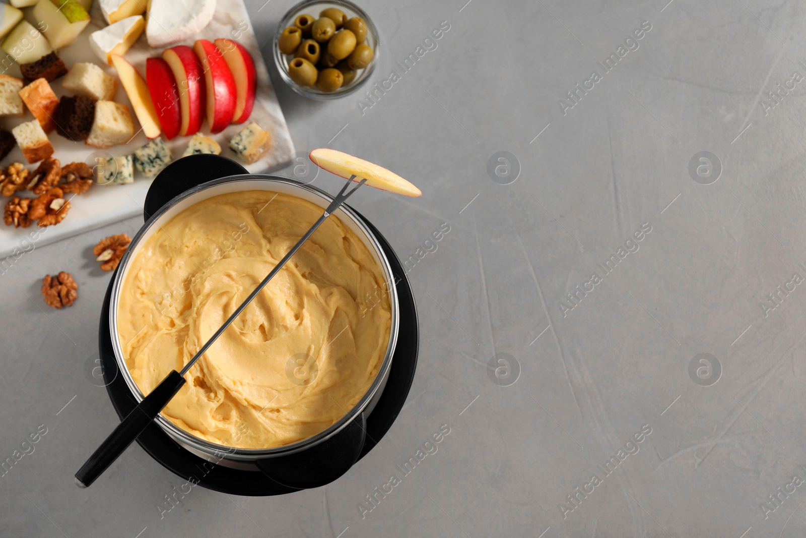 Photo of Fork with piece of apple, melted cheese in fondue pot and snacks on grey table, flat lay. Space for text