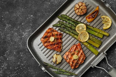 Frying pan with tasty grilled salmon, lemon and asparagus on black table, top view. Space for text