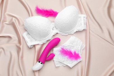 Photo of Vibrator, feathers and lingerie on beige silky fabric, flat lay