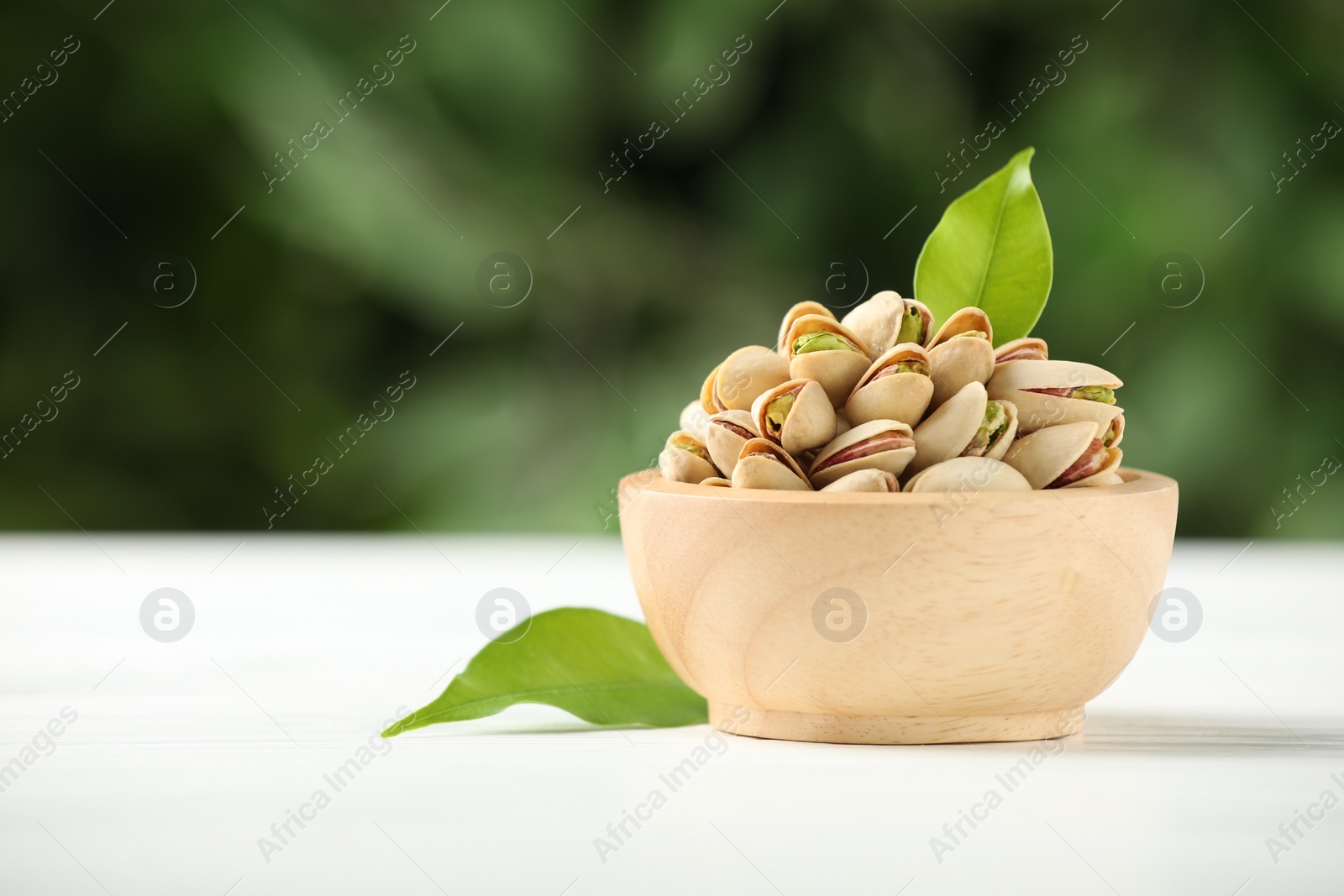 Photo of Tasty pistachios in bowl on white wooden table against blurred background, closeup. Space for text