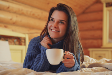 Young woman in warm sweater with cup of hot drink on bed at home
