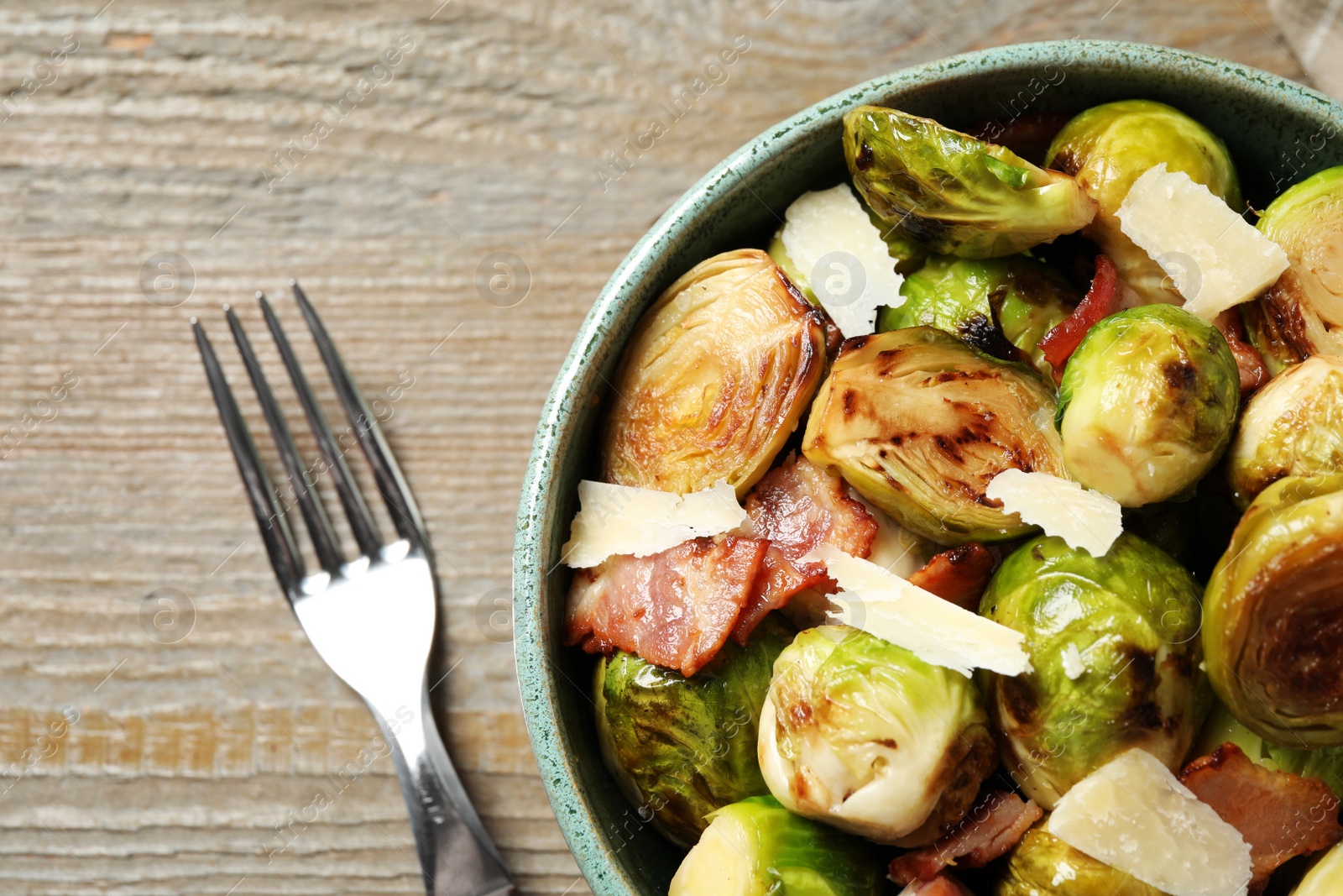 Image of Delicious fried Brussels sprouts with bacon in bowl and fork on wooden table, flat lay