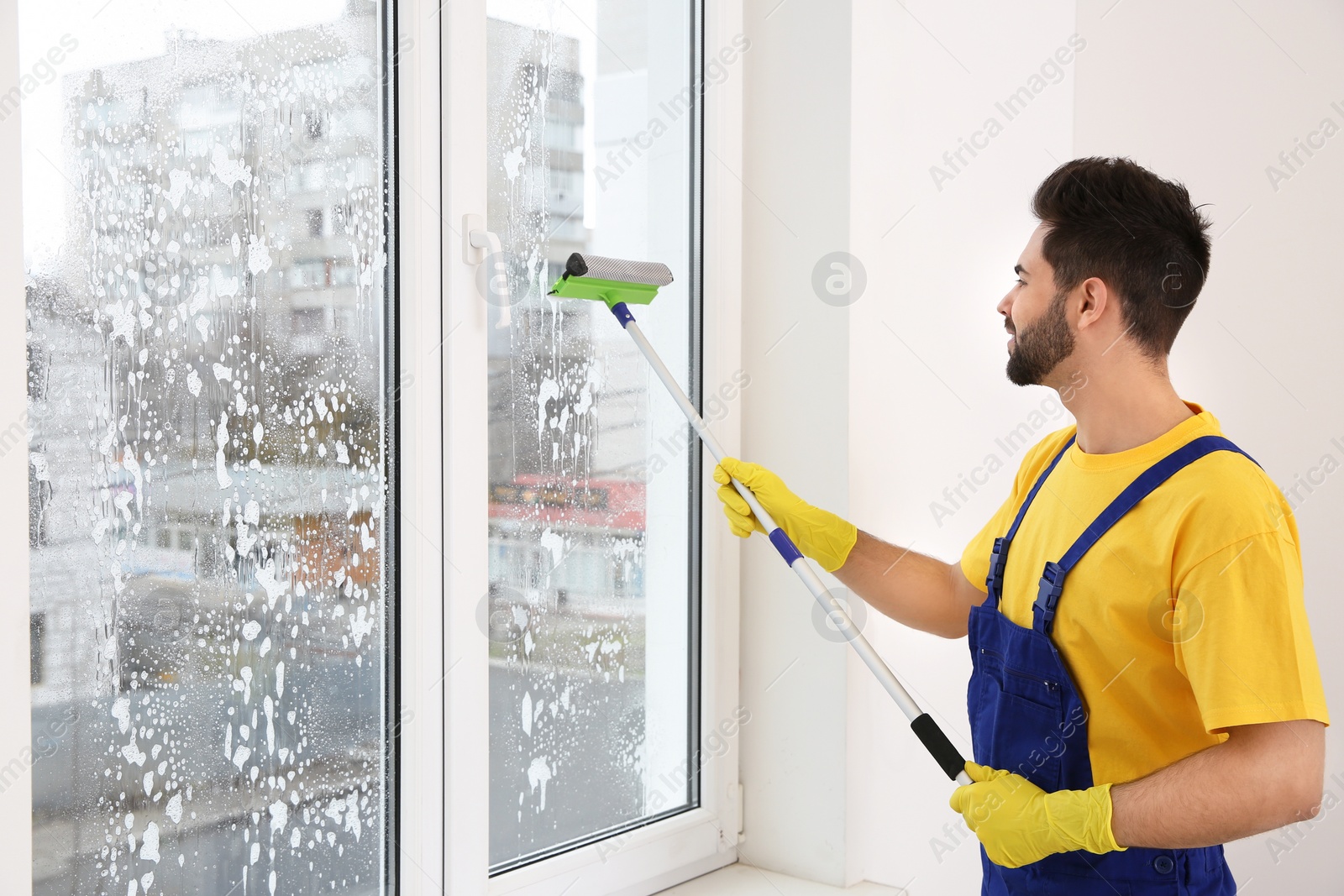 Photo of Professional young janitor in uniform cleaning window indoors