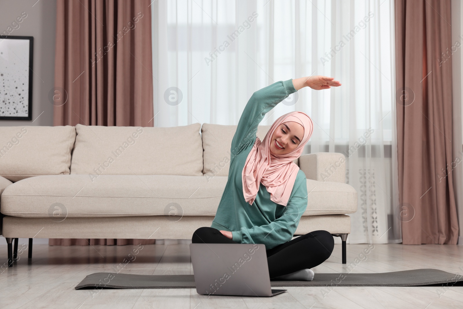 Photo of Muslim woman in hijab doing exercise near laptop on fitness mat at home. Space for text