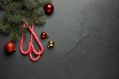 Tasty candy canes, fir branches and Christmas balls on black table, flat lay. Space for text