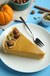 Photo of Piece of delicious pumpkin pie with hazelnuts, seeds and fork on light blue wooden table, above view