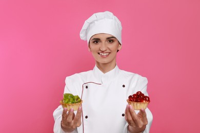 Photo of Happy professional confectioner in uniform with delicious tartlets on pink background