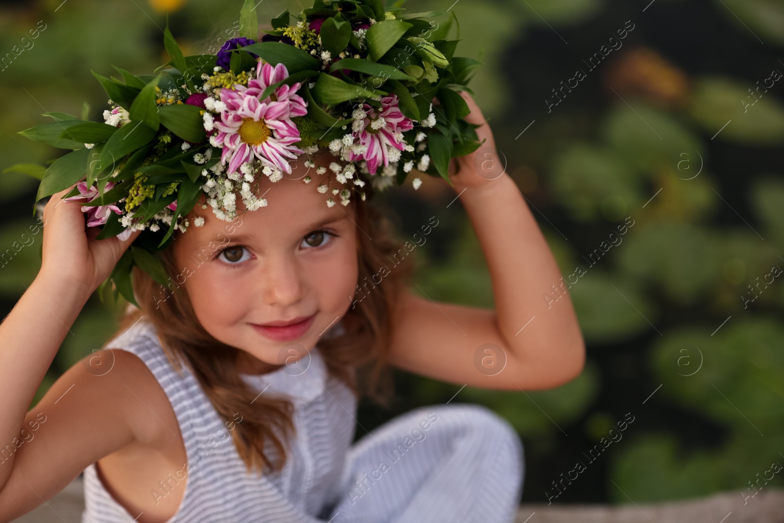 Photo of Cute little girl wearing wreath made of beautiful flowers near pond