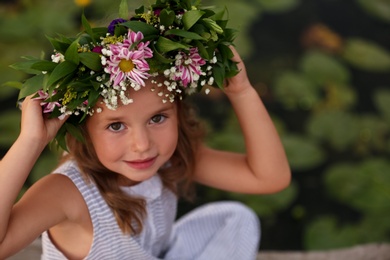 Cute little girl wearing wreath made of beautiful flowers near pond