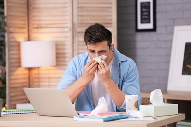 Sad exhausted man with tissue suffering from cold while working with laptop at table