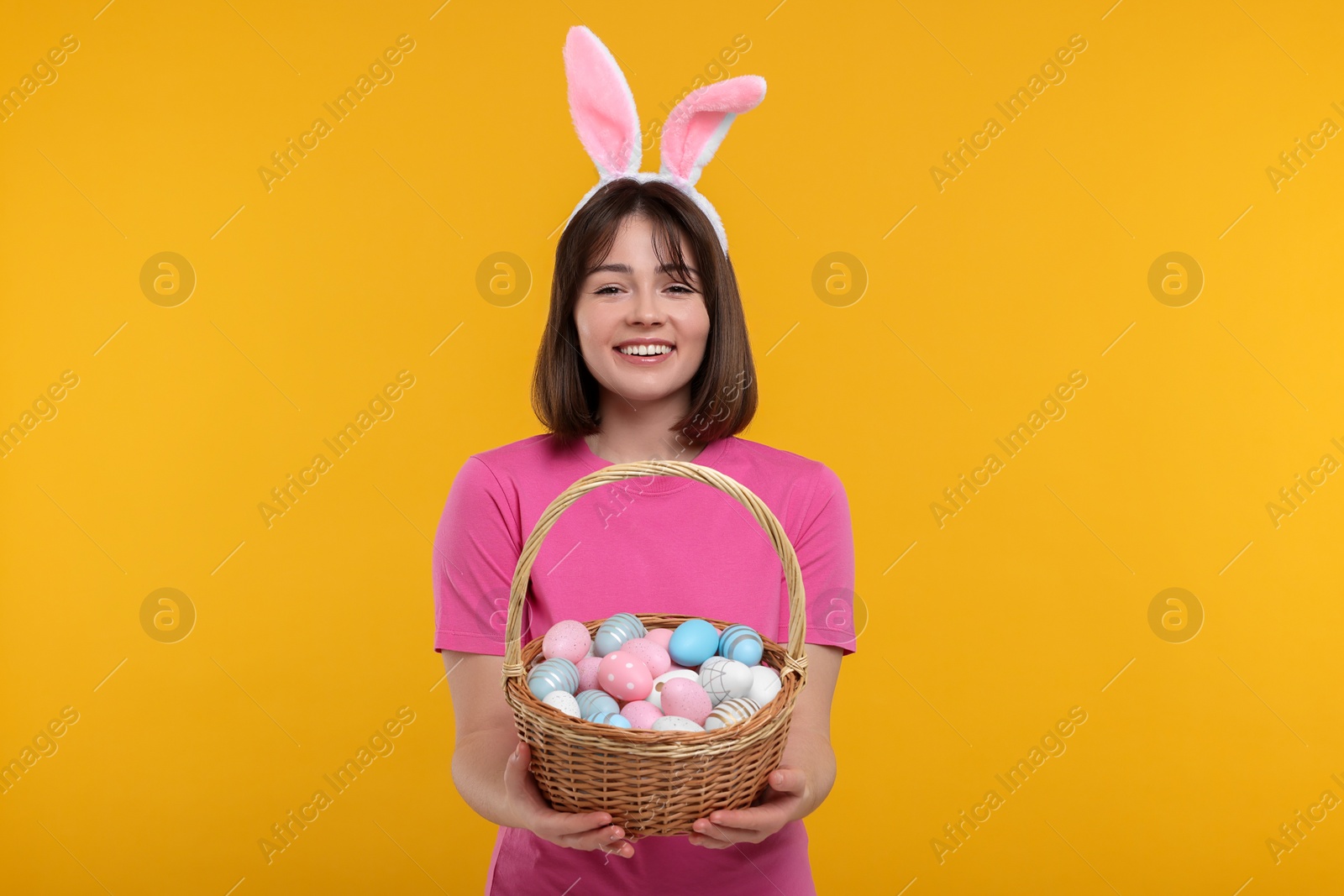 Photo of Easter celebration. Happy woman with bunny ears and wicker basket full of painted eggs on orange background