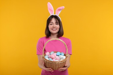 Photo of Easter celebration. Happy woman with bunny ears and wicker basket full of painted eggs on orange background