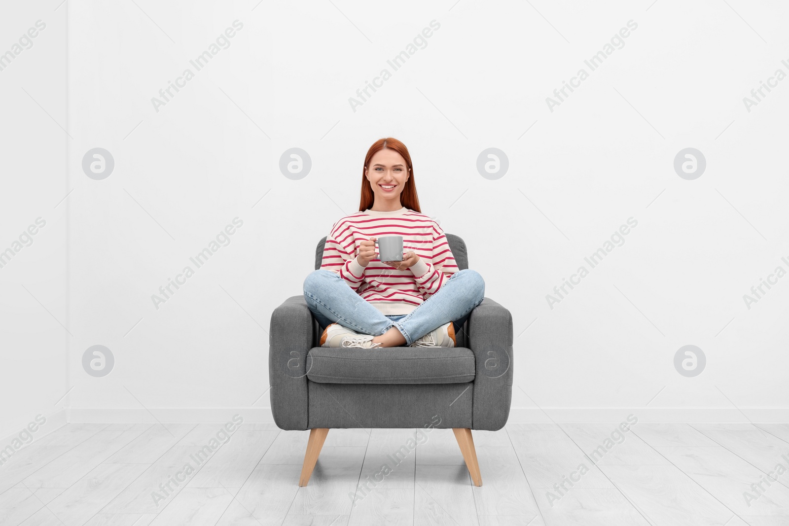 Photo of Happy young woman with cup of drink sitting in armchair near white wall indoors