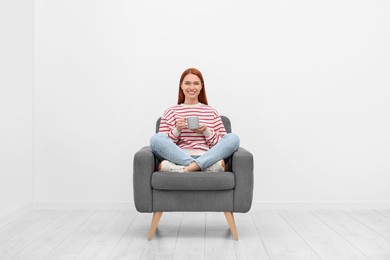 Happy young woman with cup of drink sitting in armchair near white wall indoors