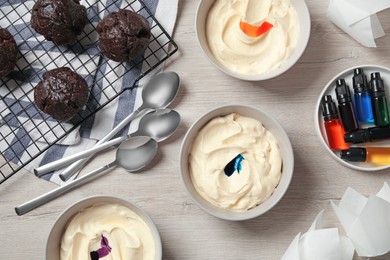 Photo of Bowls of different cream with food coloring, cupcakes, bottles of bright liquid and spoons on white wooden table, flat lay