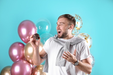 Photo of Young man with birthday muffin and air balloons on color background