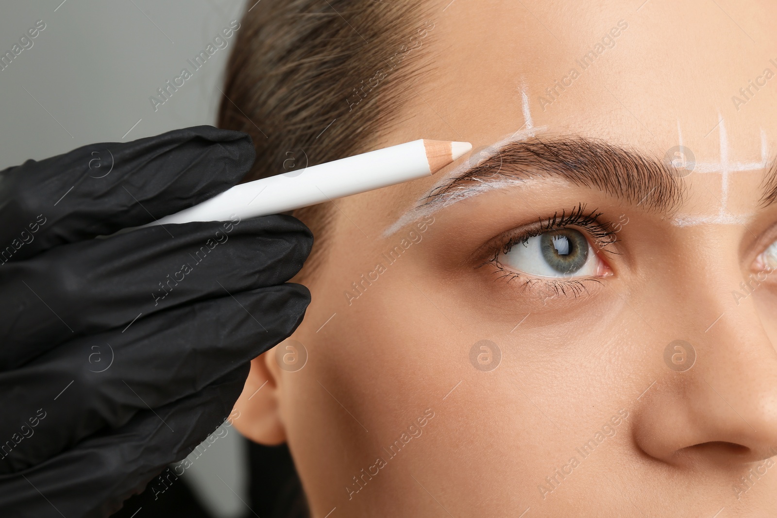 Photo of Beautician preparing young woman for procedure of permanent eyebrow makeup on grey background, closeup