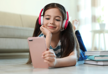 Photo of Cute little girl with headphones and smartphone listening to audiobook at home