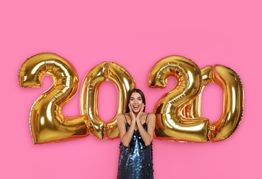 Excited young woman near golden 2020 balloons on pink background. New Year celebration