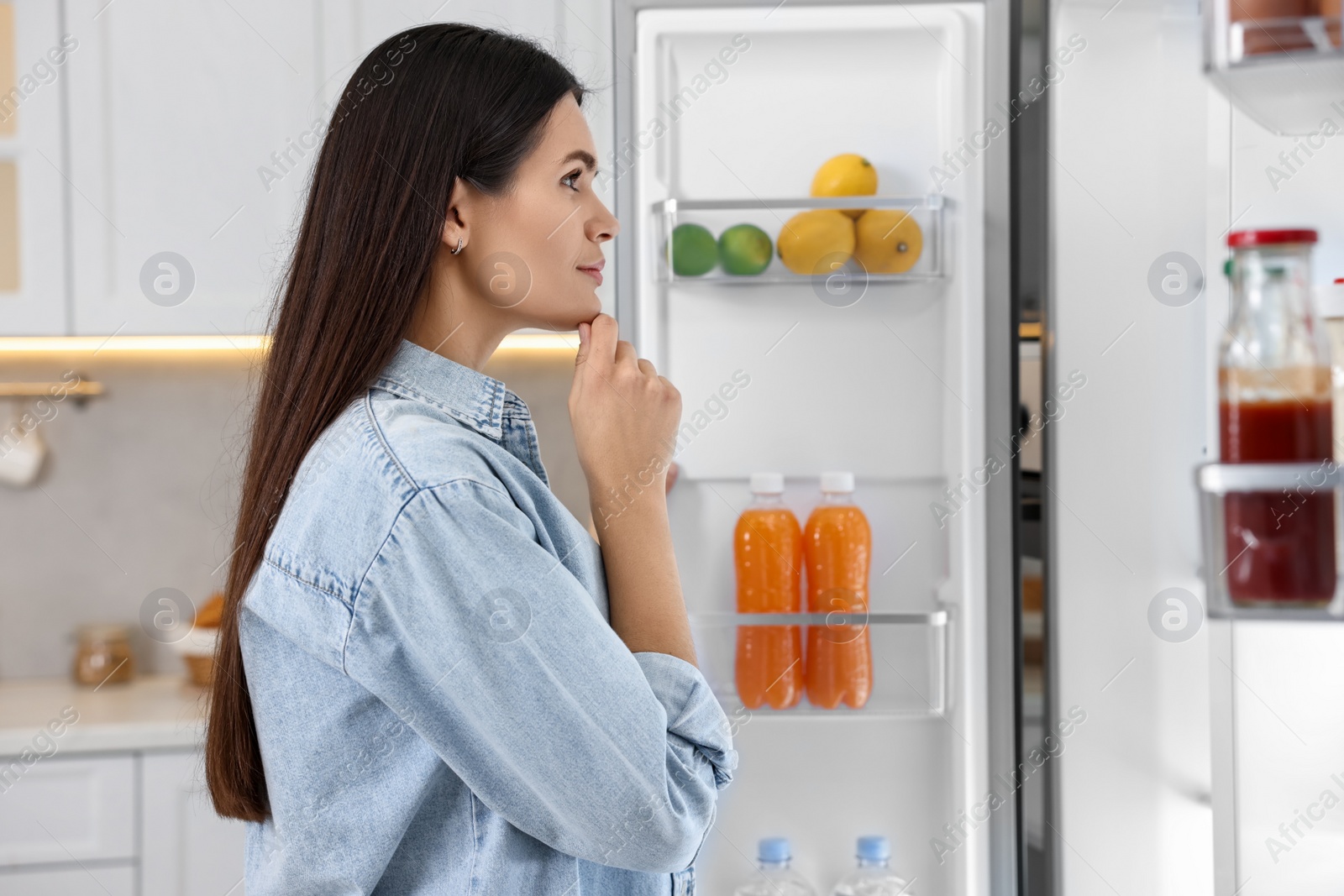 Photo of Thoughtful young woman near modern refrigerator in kitchen