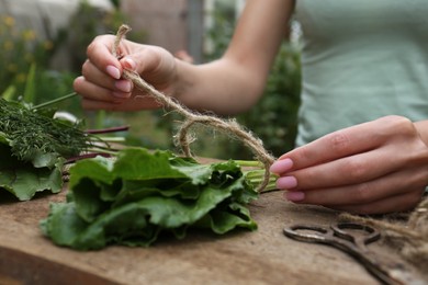 Photo of Woman tying bunch of fresh green leaves with twine at wooden table outdoors, closeup. Drying herbs