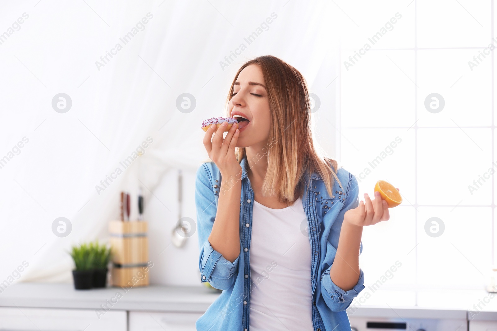 Photo of Beautiful young woman eating donut instead of orange in kitchen. Failed diet