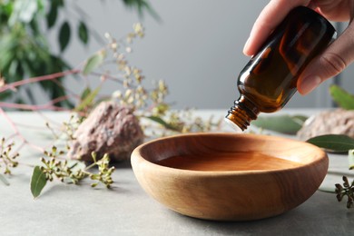 Photo of Woman dripping eucalyptus essential oil from bottle into bowl at light grey table, closeup. Space for text