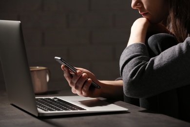 Photo of Woman using smartphone at table with laptop in dark room, closeup. Loneliness concept