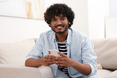 Photo of Handsome smiling man using smartphone on sofa in room