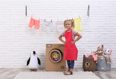Photo of Little girl playing with toy cardboard washing machine indoors