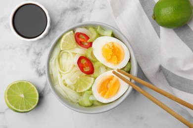 Photo of Bowl of delicious rice noodle soup with celery and egg on white marble table, flat lay