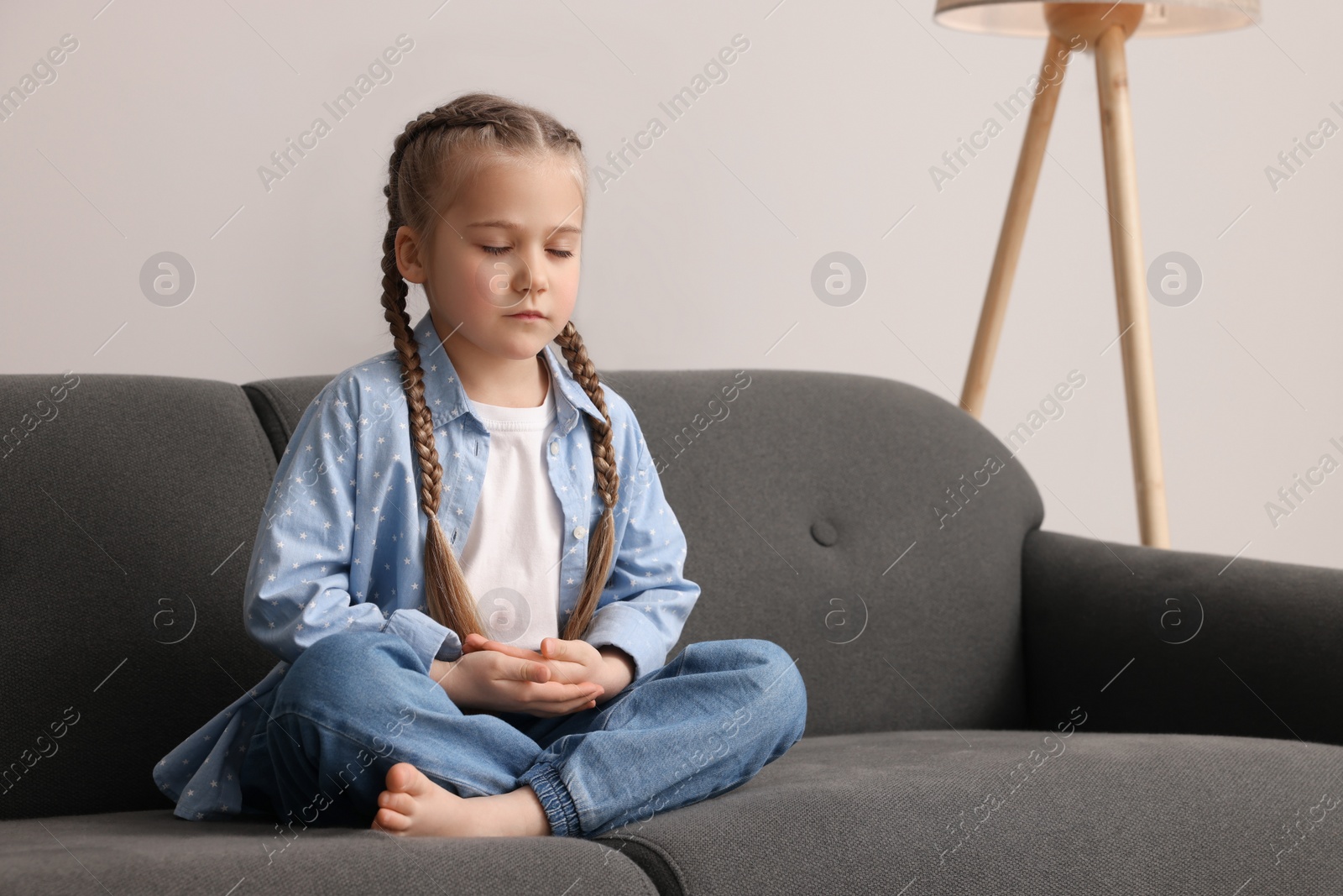 Photo of Little girl meditating on soft sofa indoors. Space for text