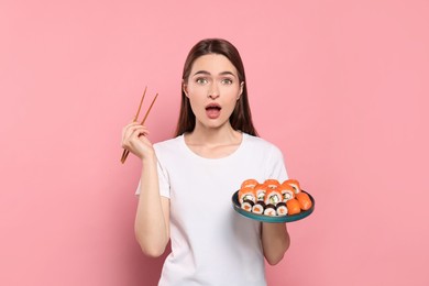 Photo of Emotional young woman with plate of sushi rolls and chopsticks on pink background