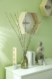 Photo of Glass vase with pussy willow tree branches and decor on white table near light green wall indoors