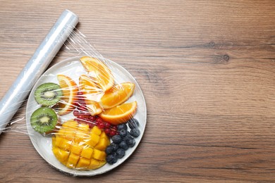 Photo of Plate of fresh fruits and berries with plastic food wrap on wooden table, flat lay. Space for text