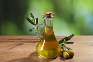 Photo of Jug of cooking oil, olives and green leaves on wooden table against blurred background