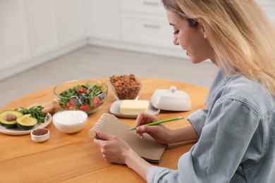 Photo of Woman with notebook and different products at wooden table in kitchen. Keto diet