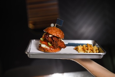 Photo of Woman holding tray with delicious burger and french fries on dark background, closeup