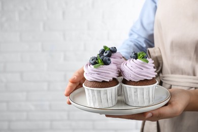 Photo of Woman holding plate with sweet cupcakes near white brick wall, closeup. Space for text