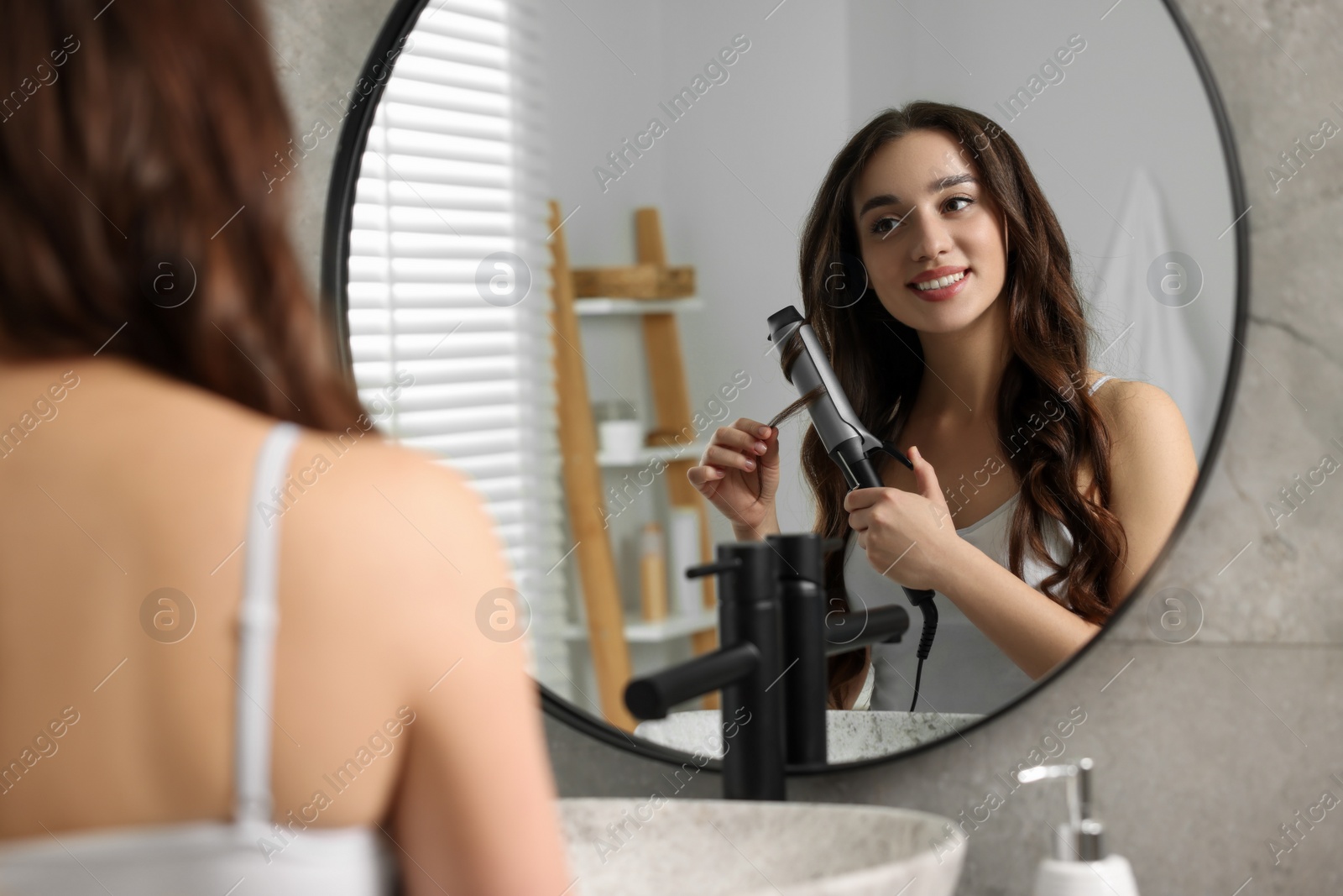 Photo of Smiling woman using curling hair iron near mirror in bathroom