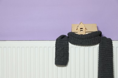 Photo of Wooden house model and knitted scarf on radiator near violet wall, space for text. Heating efficiency