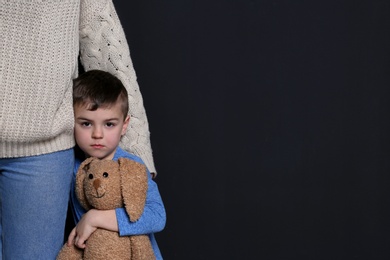 Sad little boy with toy hugging his mother on black background, space for text