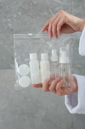 Woman holding plastic bag of cosmetic travel kit against grey wall, closeup. Bath accessories