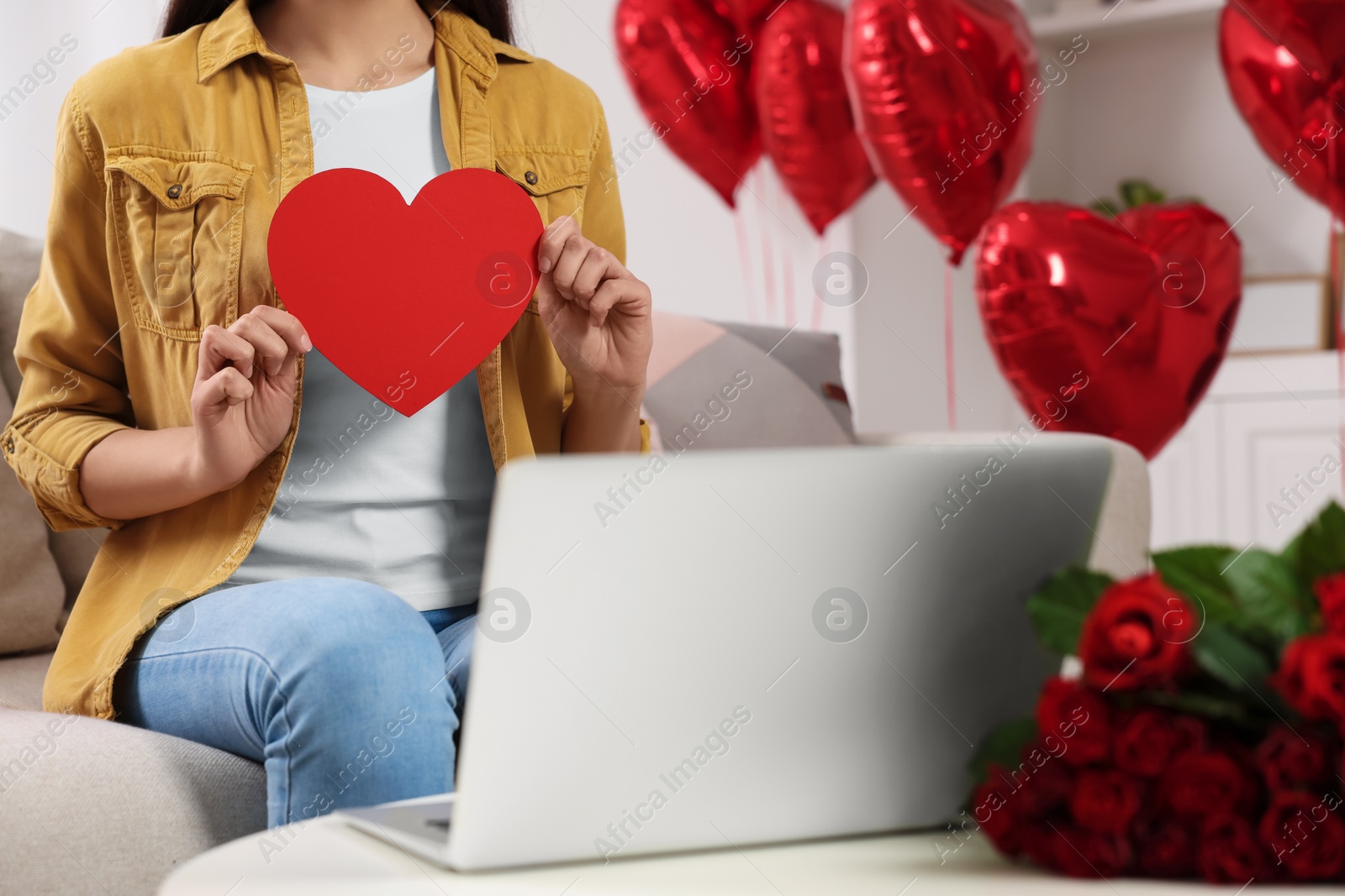 Photo of Valentine's day celebration in long distance relationship. Woman holding red paper heart while having video chat with her boyfriend via laptop, closeup