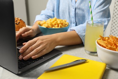 Photo of Bad eating habits. Woman using laptop surrounded by different snacks at light grey marble table, closeup