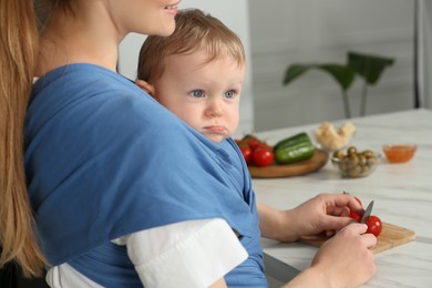 Mother cutting tomatoes while holding her child in sling (baby carrier) indoors