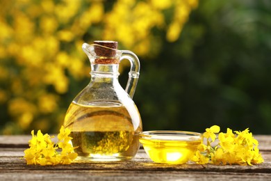 Photo of Rapeseed oil in glass jug, bowl and beautiful yellow flowers on wooden table outdoors