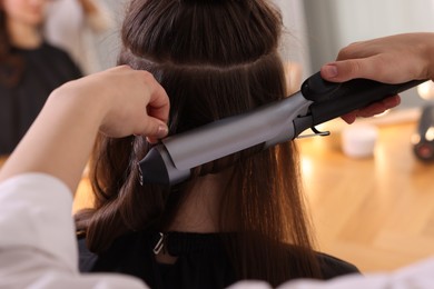 Photo of Hairdresser curling woman's hair with iron in salon, closeup